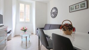 Una sala de estar moderna con una mesa de centro de vidrio, un sofá gris, una mesa de comedor blanca y una cesta de mimbre con flores.