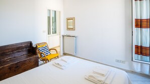 Una camera da letto con un letto, una panca di legno, una sedia con un cuscino giallo, un termosifone, uno specchio e una finestra con tende.