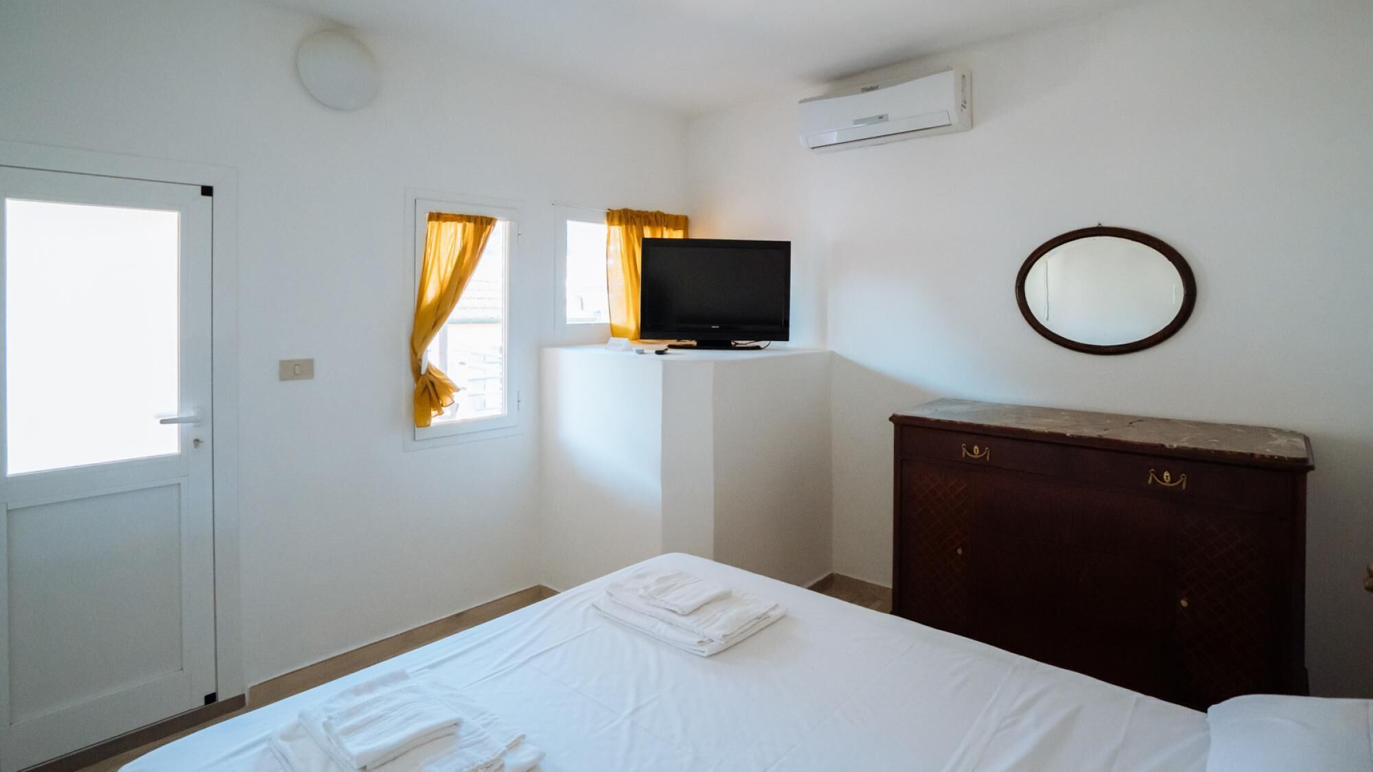 A bedroom with a bed, a television on a stand, a dresser, a mirror, and a window with curtains.