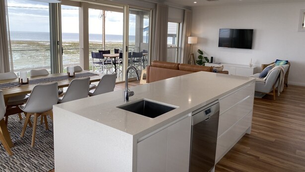A modern kitchen with a central island, a dining area with chairs and a table, a sofa, and a TV mounted on the wall.