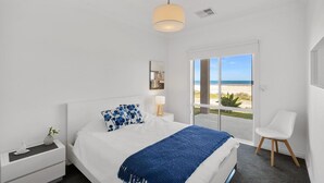 A bedroom with a bed, bedside table, lamp, chair, and a view of the beach through a glass door.