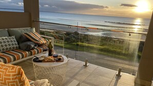 A balcony with a sofa, a round coffee table with snacks and drinks, and a view of the sea at sunset.