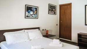 Una habitación de hotel con una cama, dos cuadros en la pared, una puerta de madera y una mesita de noche con una lámpara.