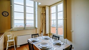 A dining area with a table set for four, large windows with curtains, a radiator, and a wall clock. - Virgilio 2 - Villa Bice (Alassio)