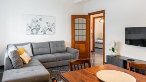 A living room with a grey sofa, a wooden dining table, a TV, and a door leading to another room.
