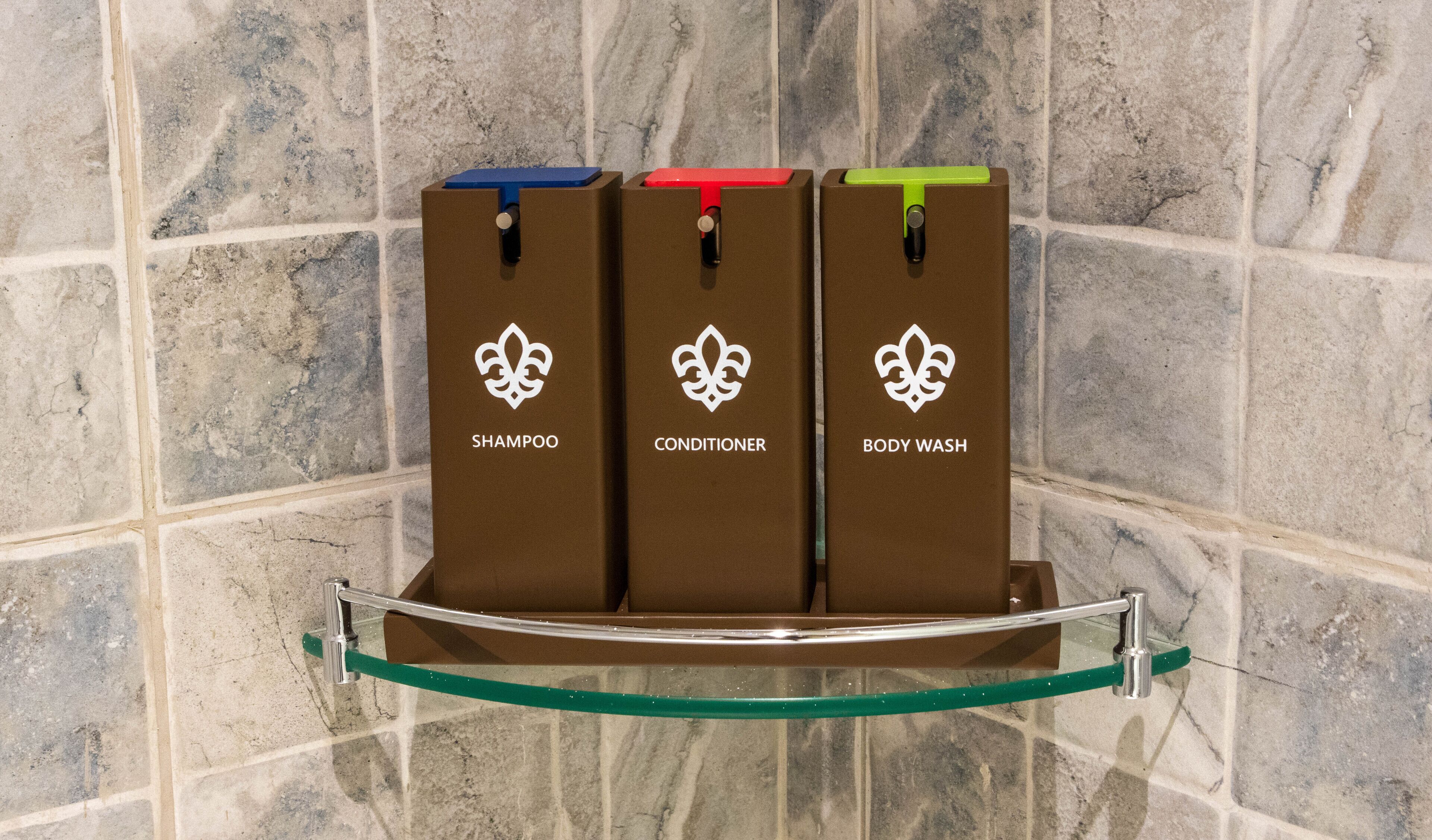 Ein Badezimmer mit Marmorwand und Ablagefläche, auf der drei braune Flaschen mit Shampoo, Conditioner und Duschgel stehen.