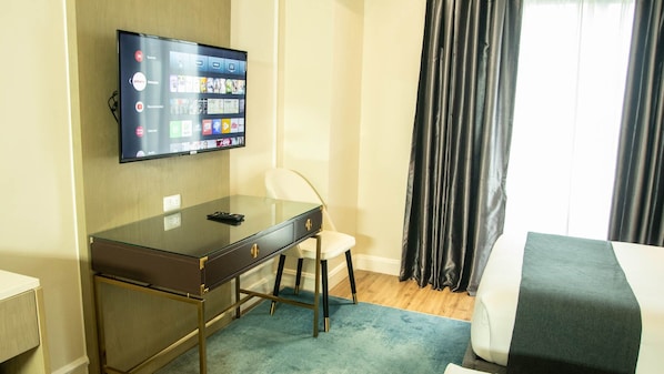 A hotel room with a TV mounted on the wall displaying various streaming services, a desk with a chair, a bed with a grey and white bedspread, and a window with black curtains. - The Noble Cebu (Cebu City)