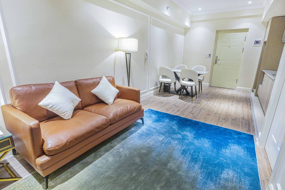 A living room with a brown leather sofa, two white cushions, a blue rug, a dining table with four chairs, and a lamp.