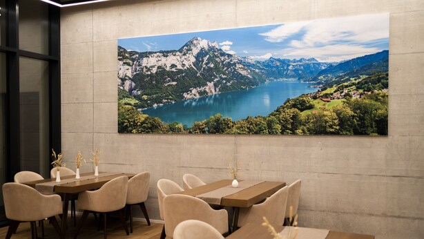 Un comedor con un gran cuadro panorámico de un paisaje montañoso y un lago, mesas de madera y sillas tapizadas en color beige.