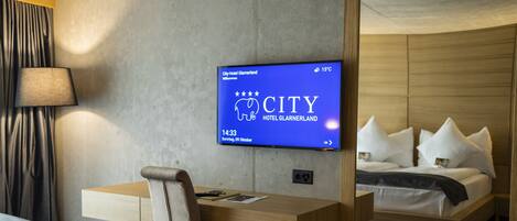 Habitación de hotel con cama, escritorio y televisor mostrando el nombre del hotel y sus servicios.