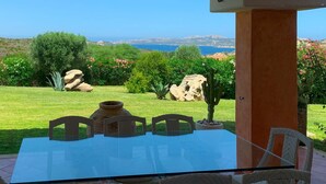 A glass-top table with four chairs, a stone pillar, and a view of a coastal landscape with greenery and rocks.