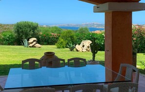 A glass-top table with four chairs, a stone pillar, and a view of a coastal landscape with greenery and rocks.
