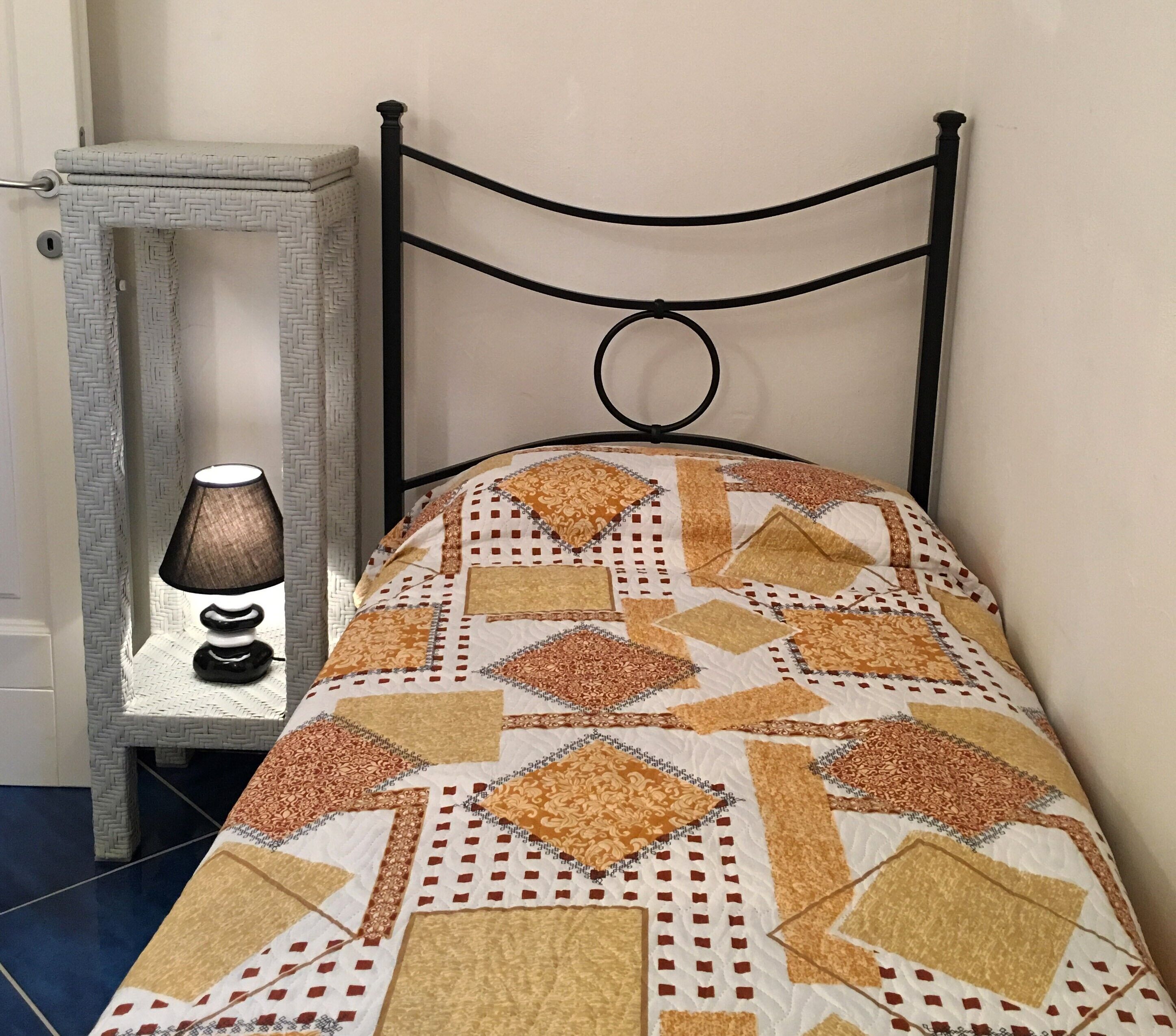 A bed with a patterned quilt, a bedside table with a lamp, and a wall-mounted headboard.