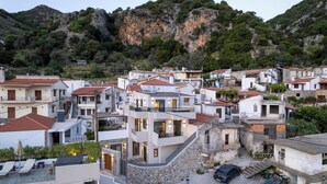 Un pueblo en una ladera con edificios modernos y tradicionales, una calle estrecha y una imponente montaña rocosa al fondo.