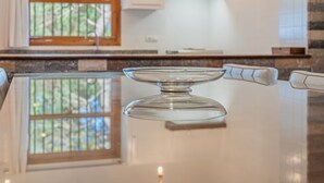 Uma tigela de vidro transparente sobre uma superfície reflexiva, uma janela com vista para árvores e uma bancada de cozinha com armários.