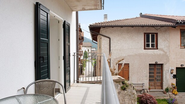 Un balcon avec des fauteuils en osier, une table en verre et une vue sur un bâtiment doté d’un toit recouvert de tuiles et d’une porte.