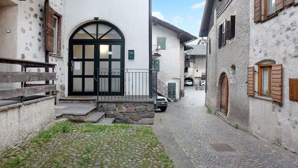 Une ruelle pavée bordée d’un bâtiment doté d’une grande entrée en arc de cercle, d’un escalier en pierre et d’un banc en bois.