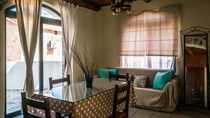 A dining area with a glass table, chairs, and a vase of branches. There is a sofa with cushions and a window with blinds. - Niriides Villas | Nisso | Villa 2-bedrooms with private pool (CHERSONISSOS-HERAKLION)
