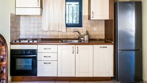 A kitchen with white cabinets, a stainless steel refrigerator, a window with a curtain, and a built-in oven. - Niriides Villas | Nisso | Villa 2-bedrooms with private pool (CHERSONISSOS-HERAKLION)