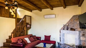 A cozy living room with a wooden staircase, a stone fireplace, and a red sofa. - Niriides Villas | Nisso | Villa 2-bedrooms with private pool (CHERSONISSOS-HERAKLION)