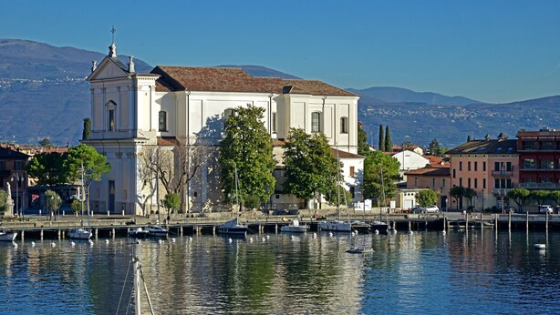 A waterfront building with a prominent bell tower, surrounded by trees and a marina with boats.