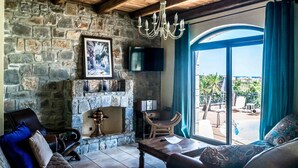 A stone-walled living room with a fireplace, a wooden ceiling, a chandelier, a sofa, and a view of an outdoor patio. - Niriides Villas | Liagori | Villa 2-bedrooms with private pool (CHERSONISSOS-HERAKLION)