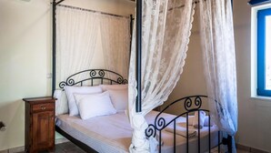 A bed with a canopy, a bedside table, and a window with blue trim. - Niriides Villas | Liagori | Villa 2-bedrooms with private pool (CHERSONISSOS-HERAKLION)