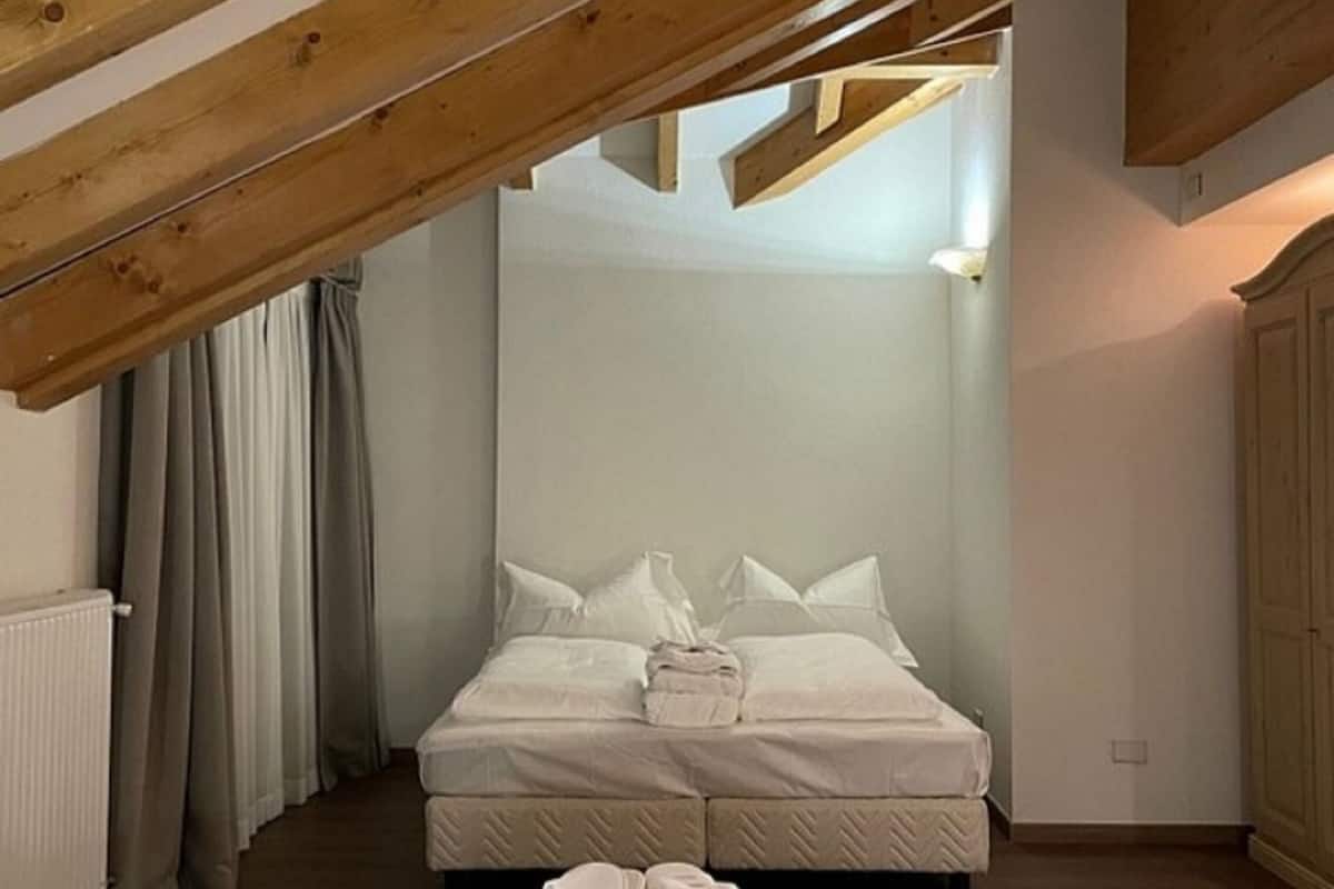 Ein Schlafzimmer mit schrägverdeckter Decke, einem Bett mit weißen Leinen, einem Heizkörper und einem Vorhangpaar.