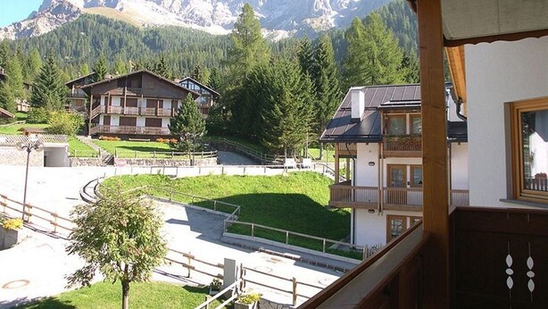 Vista de la montaña con un edificio de estilo chalet y una casa más pequeña con balcón, todo rodeado de vegetación y una zona pavimentada.
