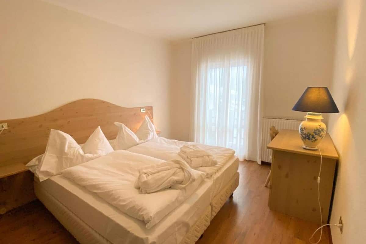 Ein ordentlich bezogenes Bett in einem Zimmer mit Holzboden, einem Nachttisch mit Lampe und einem Fenster mit Vorhängen.