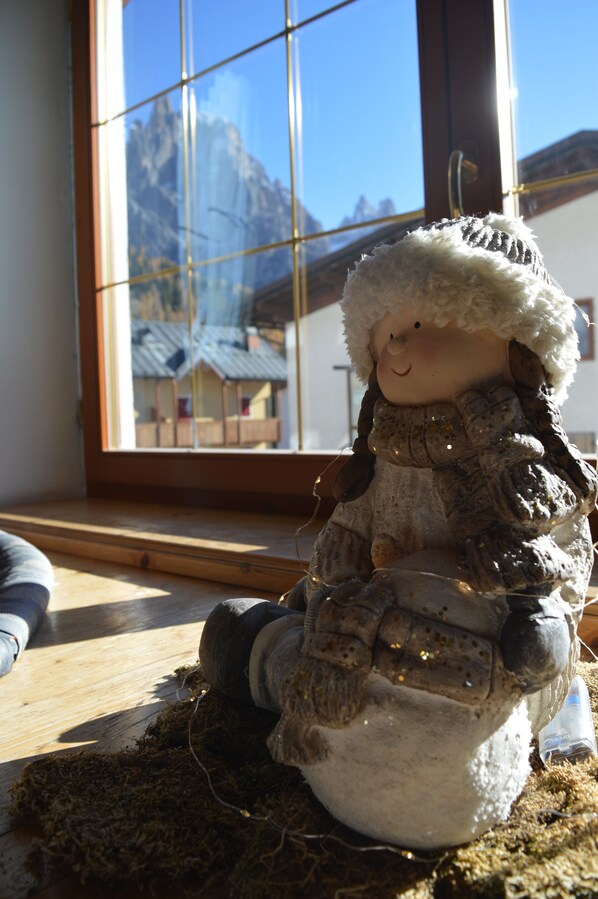 Une poupée portant un chapeau et un manteau blancs, assise sur une surface en bois près d’une fenêtre donnant sur une vue de montagne.