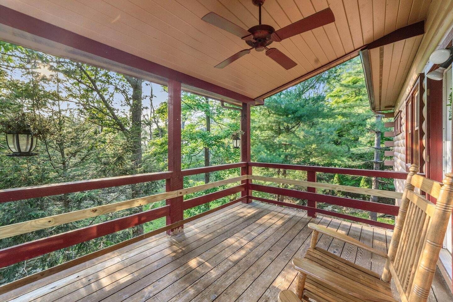 A wooden porch with a red railing, a ceiling fan, and a view of a forested area.