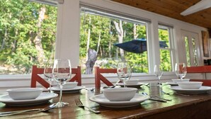 Une table à manger dressée avec de l’assiette blanche, des bols et des verres à vin, donnant sur un paysage arboré grâce à de grandes fenêtres.