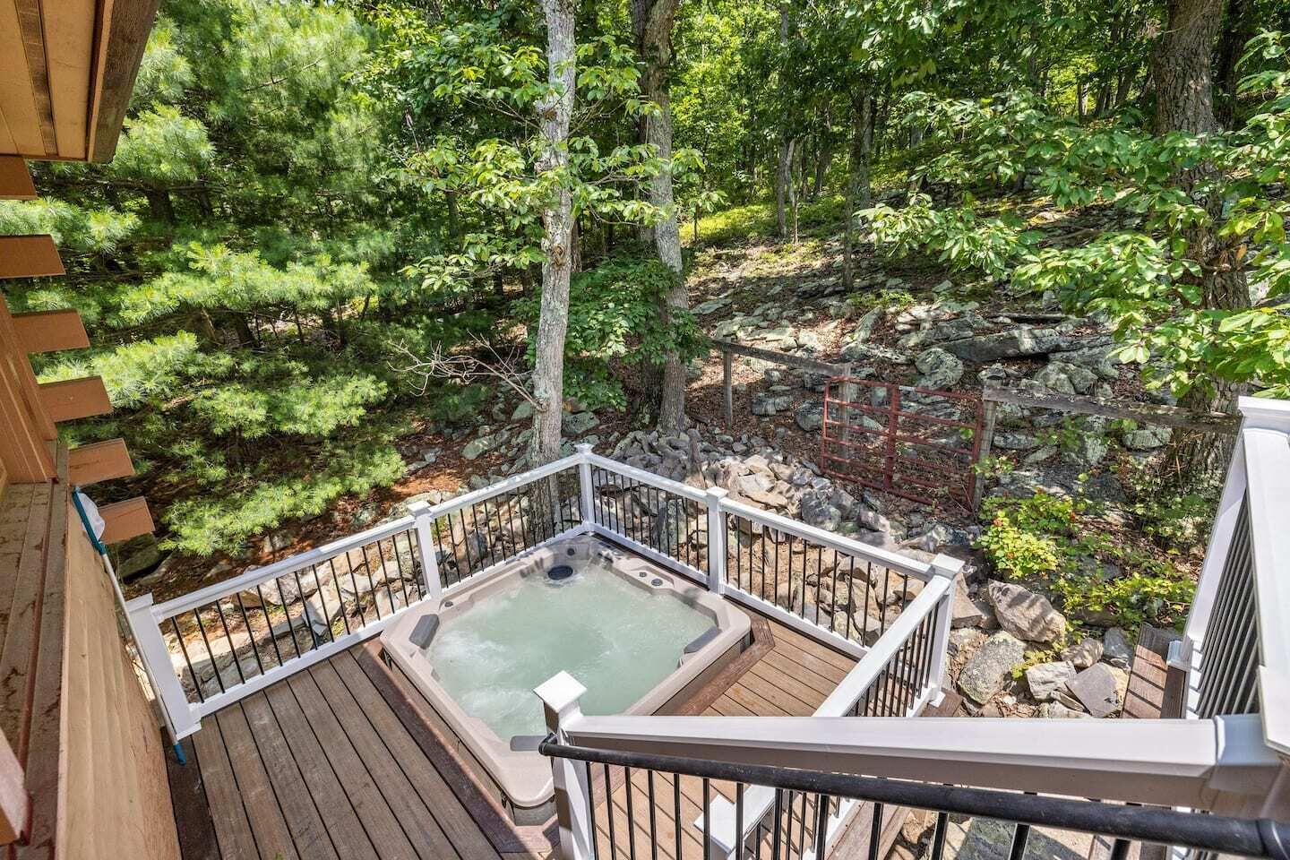 Un bain à remous sur une terrasse en bois, entouré d’une balustrade blanche, avec vue sur une zone boisée parsemée de rochers et d’arbres.