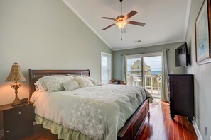 A bedroom with a bed, a nightstand, a lamp, a ceiling fan, a TV, and a balcony with a view of the sea.
