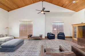 A spacious living room with a high ceiling, a ceiling fan, two windows with blinds, a sofa, a chair, and a cabinet.
