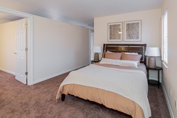 A bedroom with a large bed, two bedside tables with lamps, a door leading to another room, and two framed pictures on the wall.