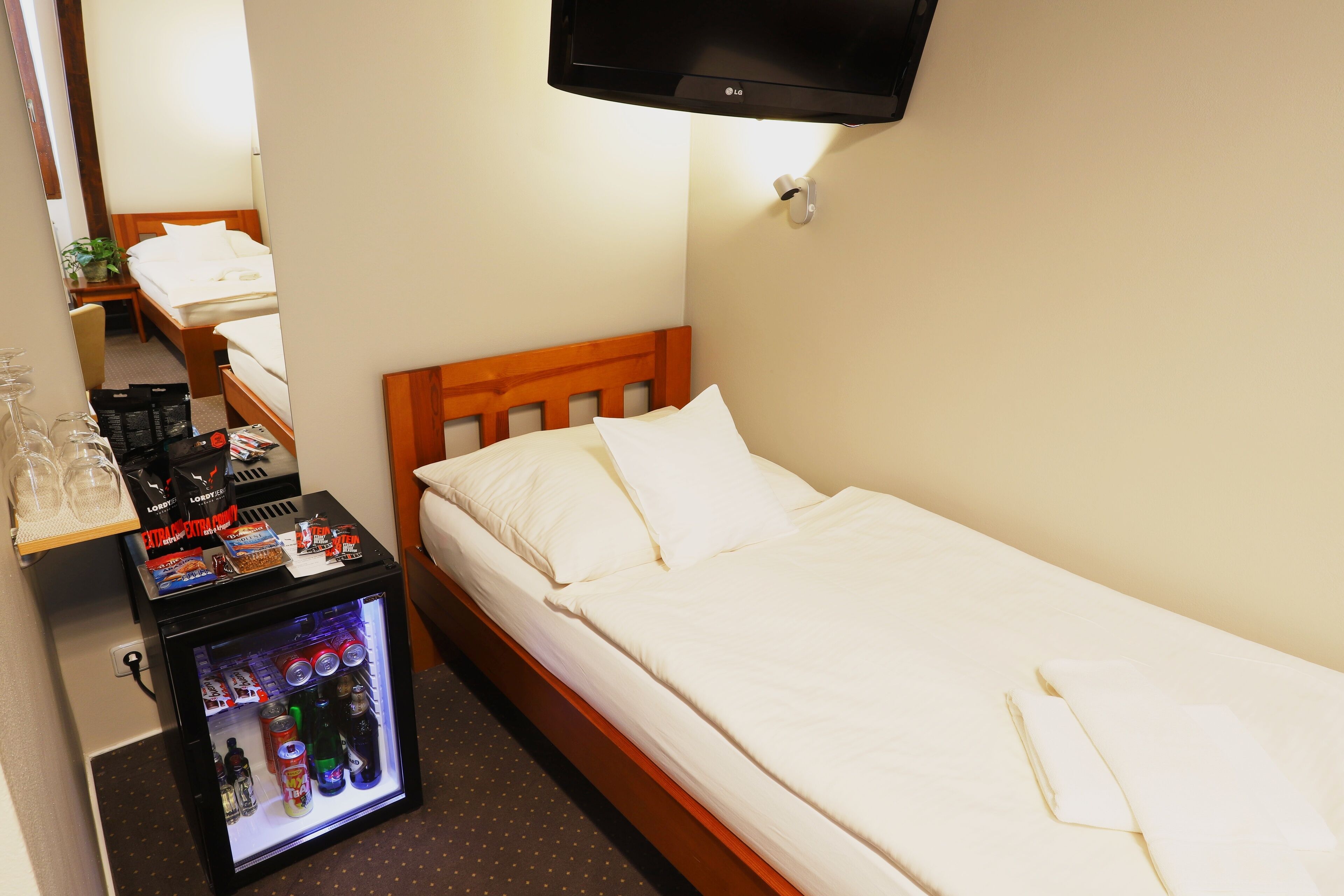 A hotel room with a bed, a small refrigerator with beverages, a television, and a bedside table with a plant and a bag.