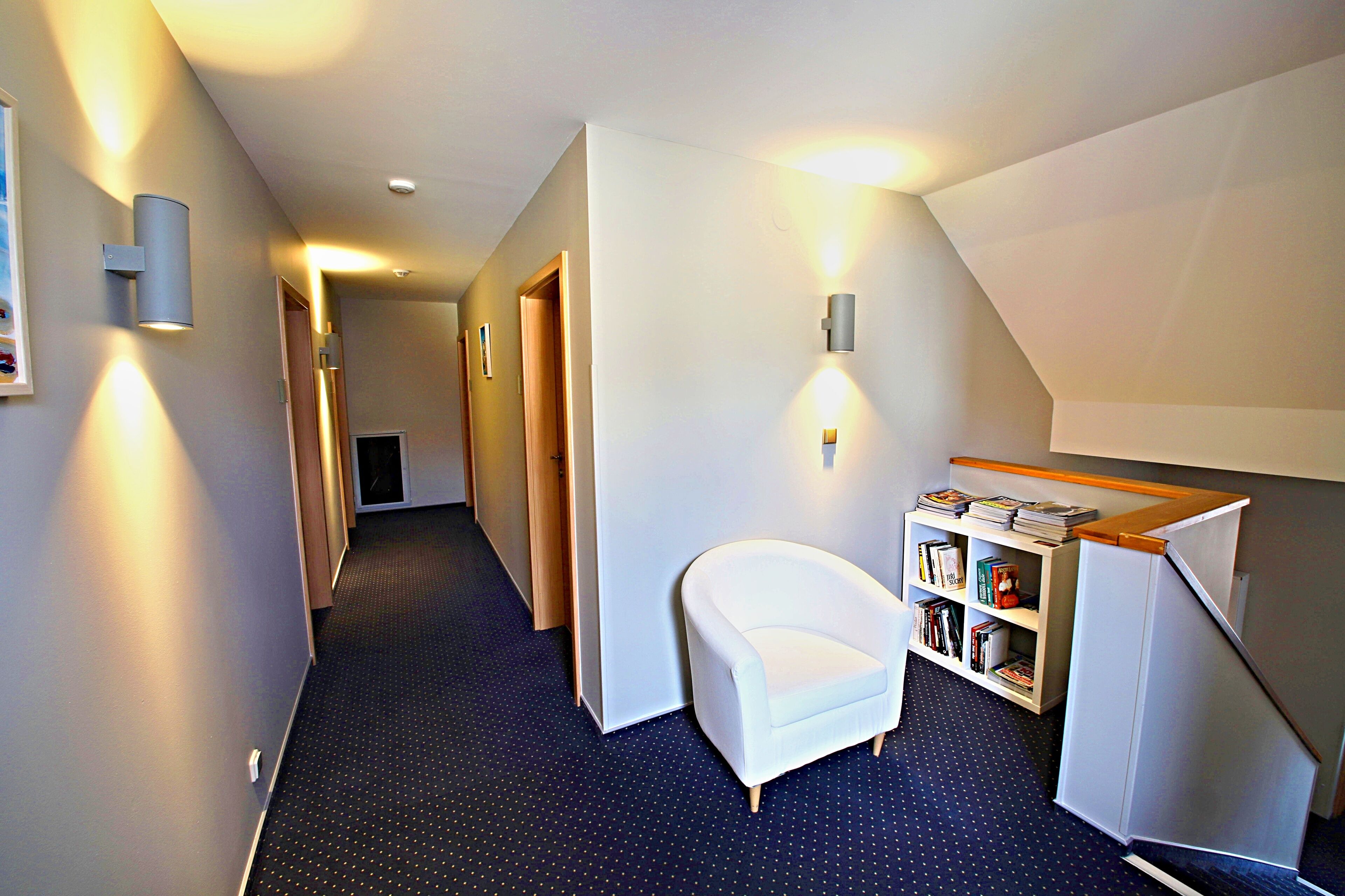 A hallway with a white chair, a bookshelf, and a staircase.