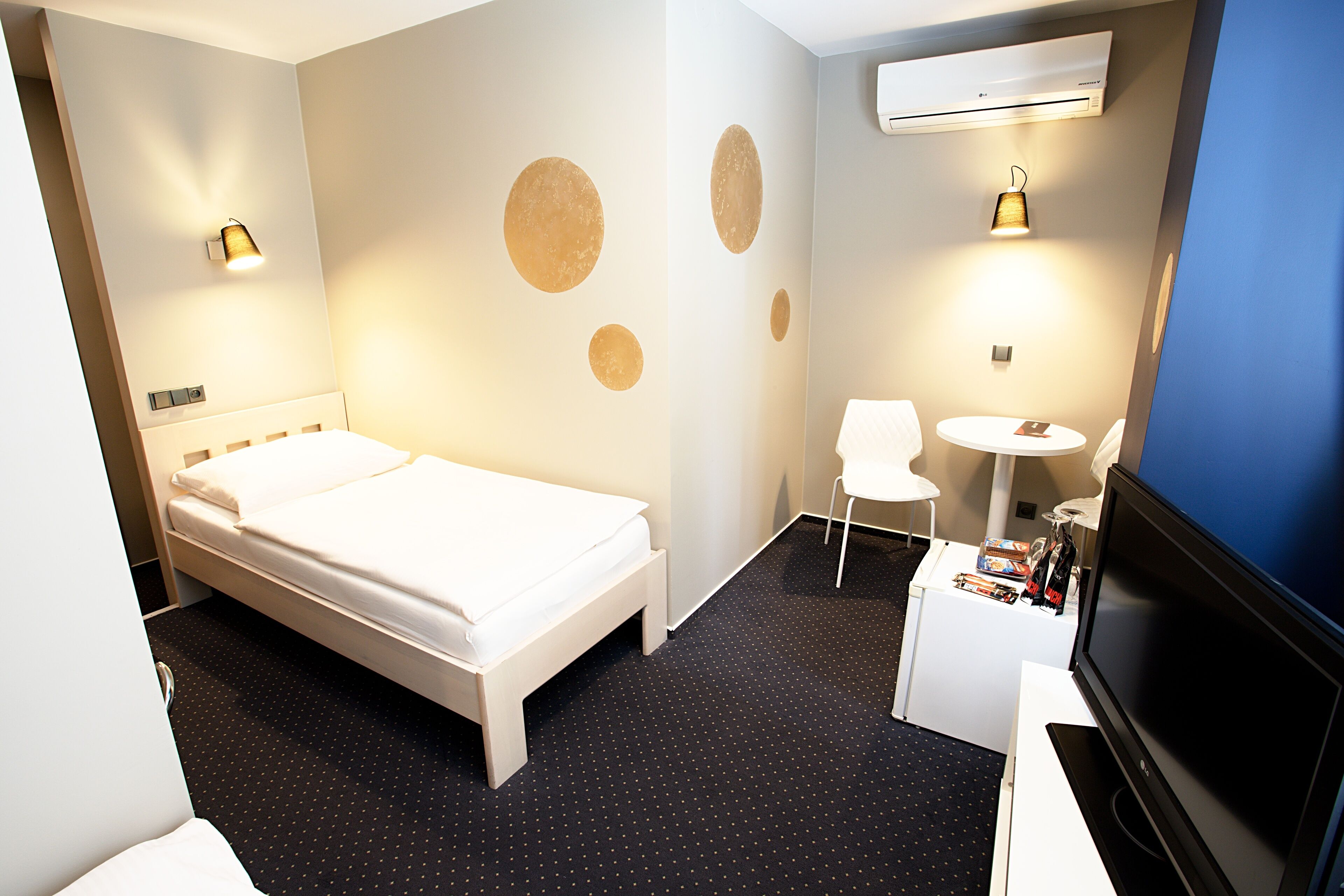 A hotel room with a bed, a desk with a chair, a TV, and wall-mounted lights.