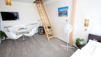 A modern hotel room with a white bed, a small table with chairs, a TV mounted on the wall, a ladder, a fan, and a painting.