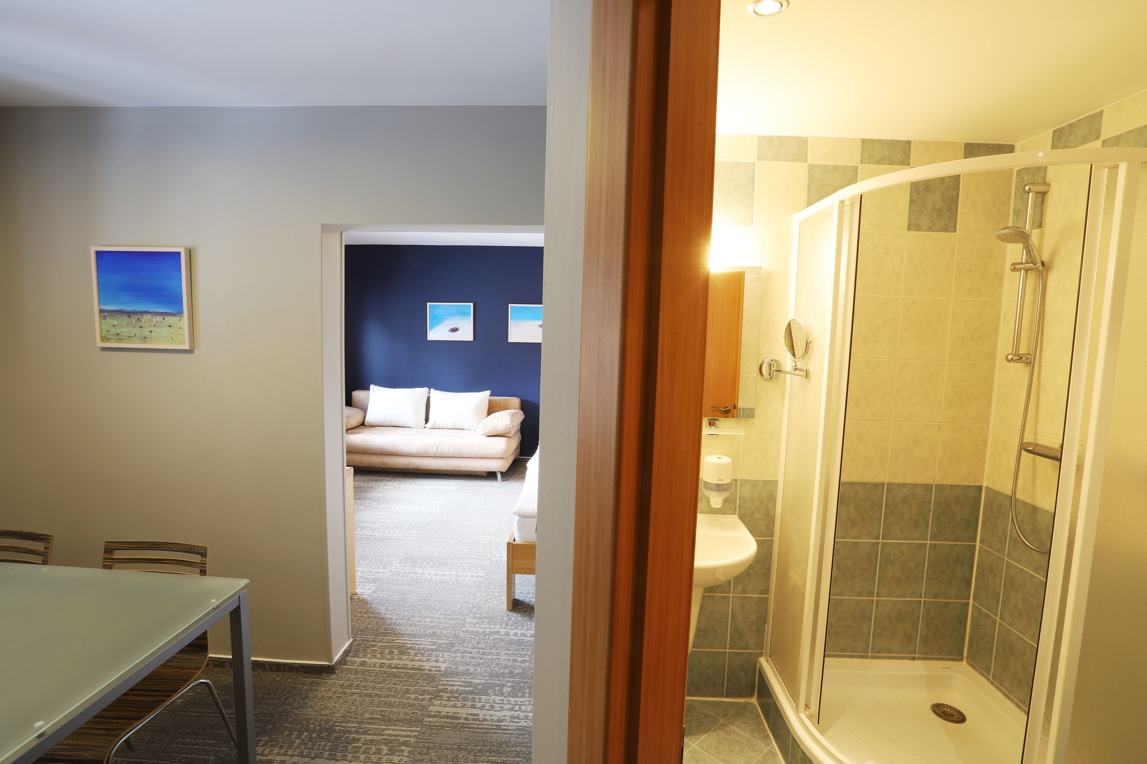 A hotel room with a glass shower, a dining area with a table and chairs, a sofa, and a view of the living area.