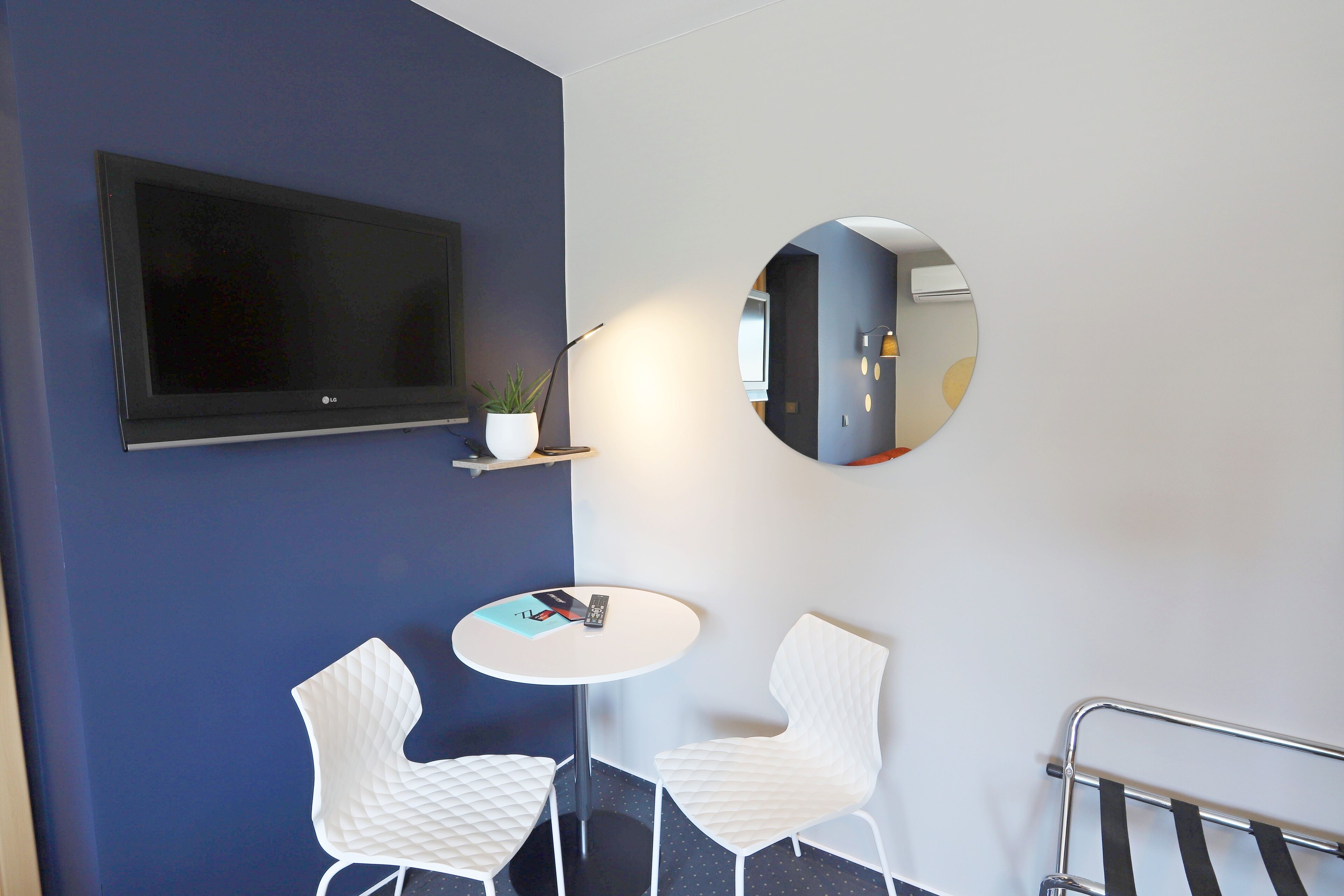 A modern room with a flat-screen TV, two white chairs around a small table, a potted plant, and a round mirror on the wall.