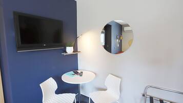 A modern room with a flat-screen TV, two white chairs around a small table, a potted plant, and a round mirror on the wall.