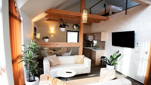 Una sala de estar moderna con un sofá blanco, una mesa de centro y un televisor montado en la pared. Se ven plantas en macetas y una zona de cocina al fondo.