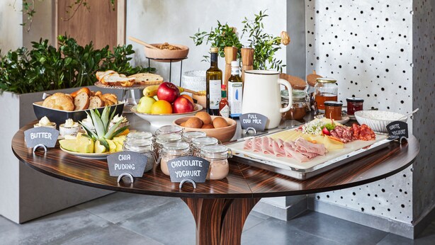 Una mesa de buffet con diversos alimentos y condimentos, incluyendo frascos etiquetados de mantequilla, pudín de chía, chocolate y yogur.