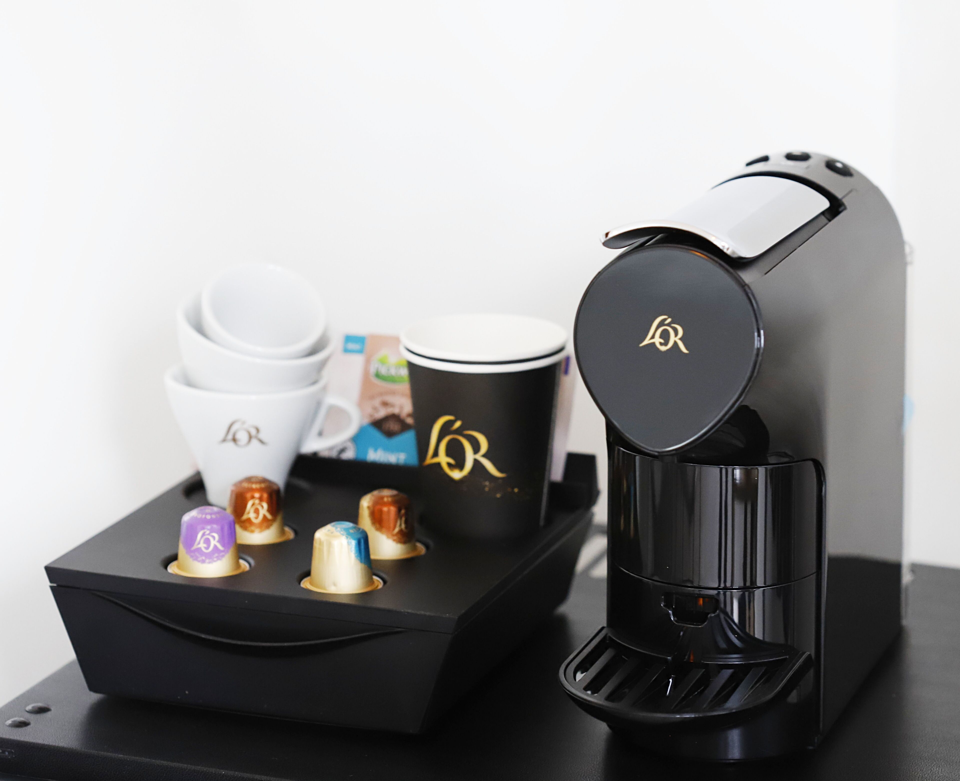 A coffee maker with a black base and a glossy top, accompanied by a black tray with coffee cups and a box of coffee pods.