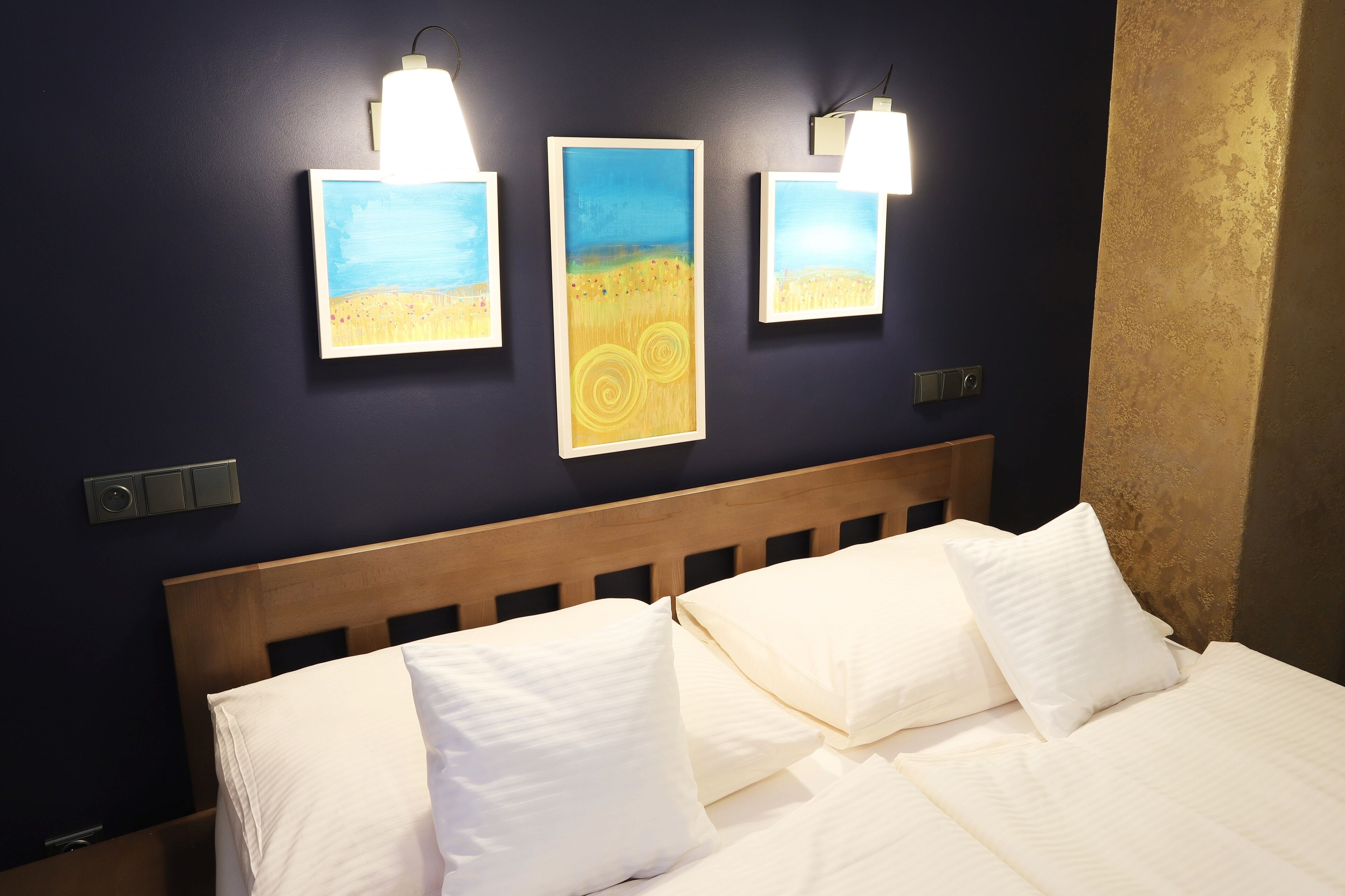 A hotel room with a wooden headboard, white bedding, and three wall-mounted paintings depicting landscapes.