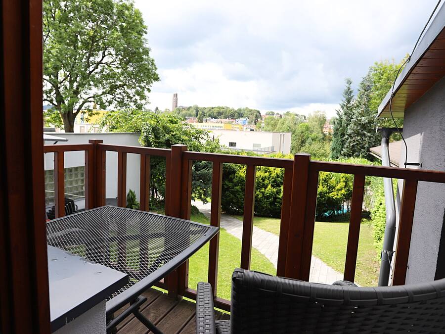 Un balcón con mesa de metal y silla de mimbre, con vistas a un jardín y edificios.