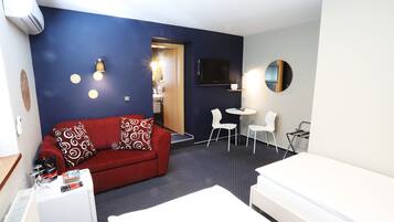 A compact hotel room with a red sofa, a white bed, a small table, and a TV mounted on the wall.